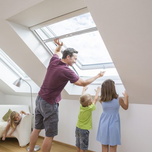Rollo für Dachfenster mit Insektenschutz, Familie lüftet gemeinsam am geöffneten Dachfenster