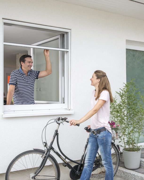 Insektenschutzrollo für Fenster, Mann öffnet das Fenster mit heruntergelassenem Fliegengitter und spricht mit Frau draußen